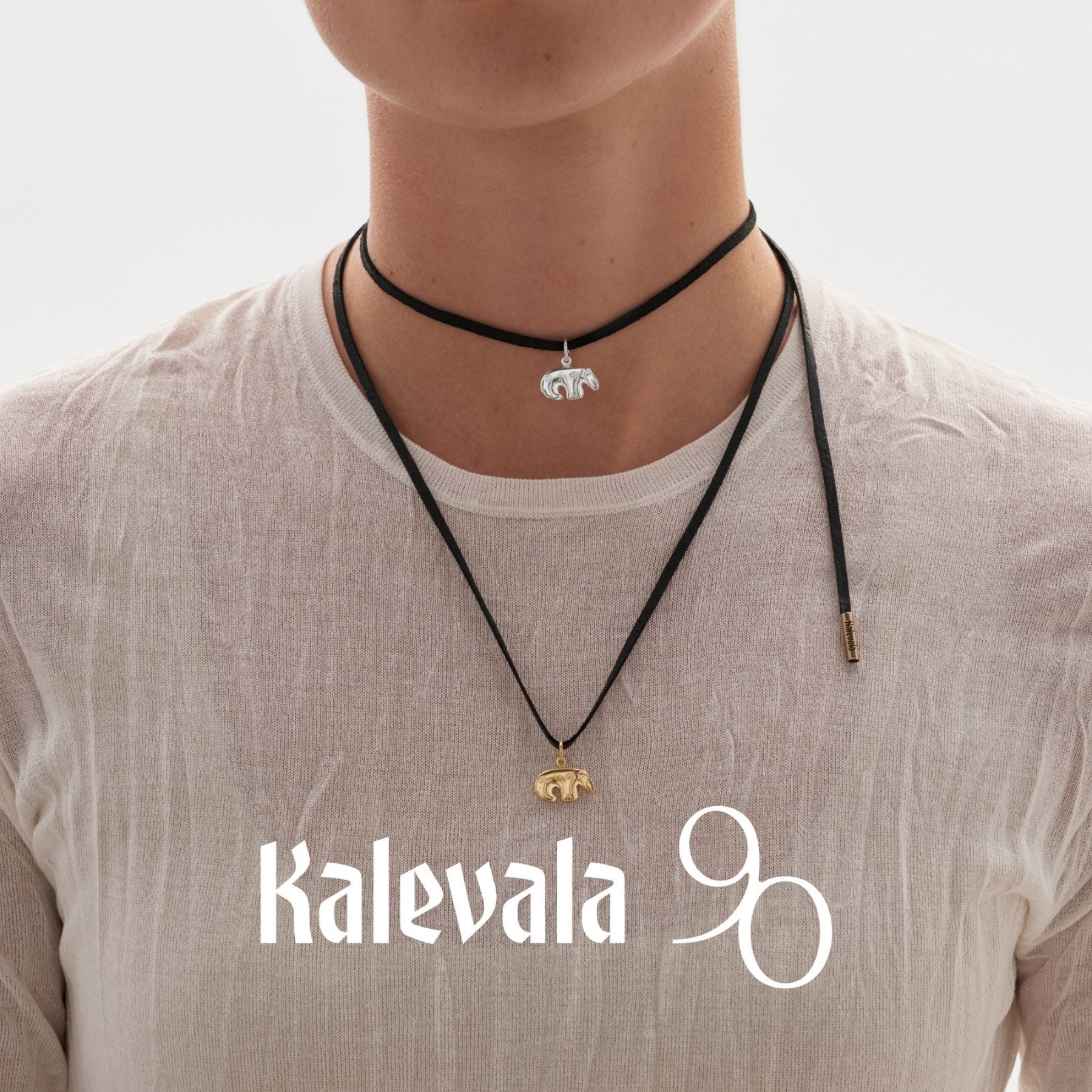 Kalevalan lämminsävyinen pronssinen karhuhela mustassa kaulanauhassa, mallina valkoiseen puseroon pukeutunut henkilö ja Kalevala 90 -tunnus