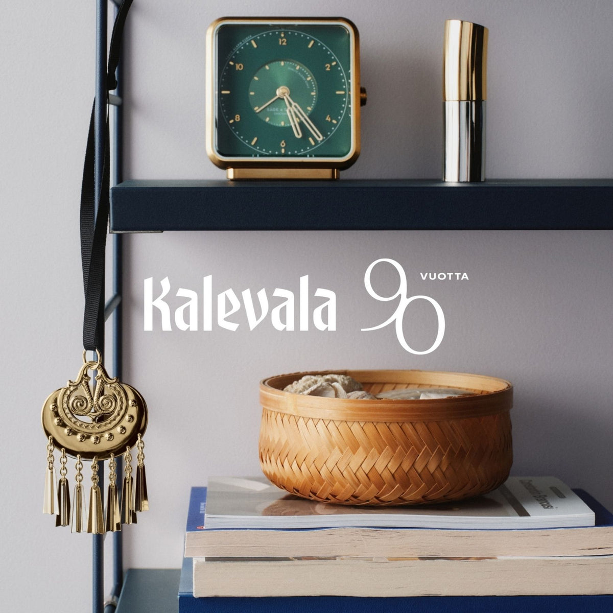 Kalevalan kullanhohtoinen pronssikoriste mustalla satiininauhalla hyllyssä, taustalla Kalevalan 90-vuotisjuhlan teksti
