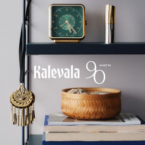 Kalevalan kullanhohtoinen pronssikoriste mustalla satiininauhalla hyllyssä, taustalla Kalevalan 90-vuotisjuhlan teksti