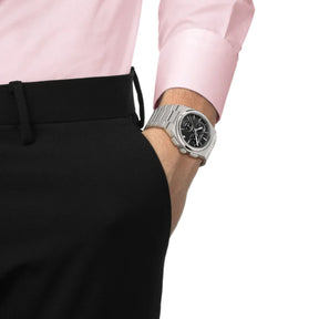 Tissot PRX kronografi teräsrannekkeella ja mustalla taululla ranteessa vaaleanpunaisen paidan kanssa